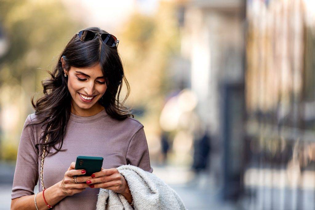 Beautiful mid adult woman walking and texting message on mobile phone outside business center.